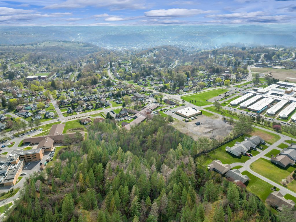Aerial view of a forested area next to a cleared plot, residential and commercial buildings. Hills in the distance.