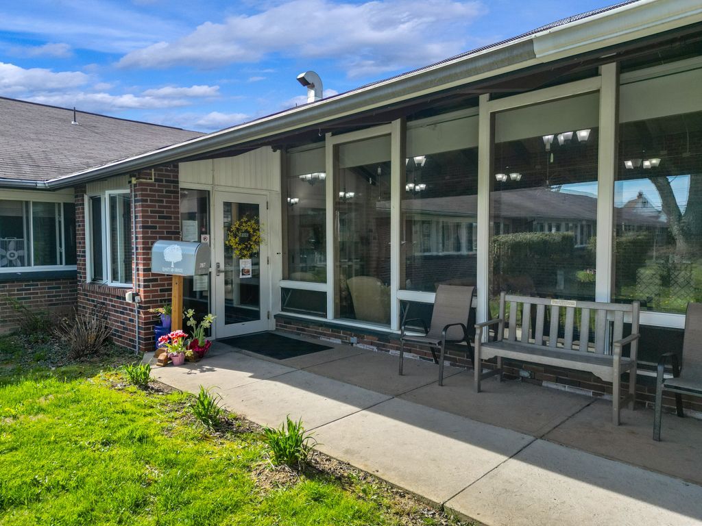 Exterior of a building with glass windows, a porch, and a bench; a mailbox and flowers are visible.