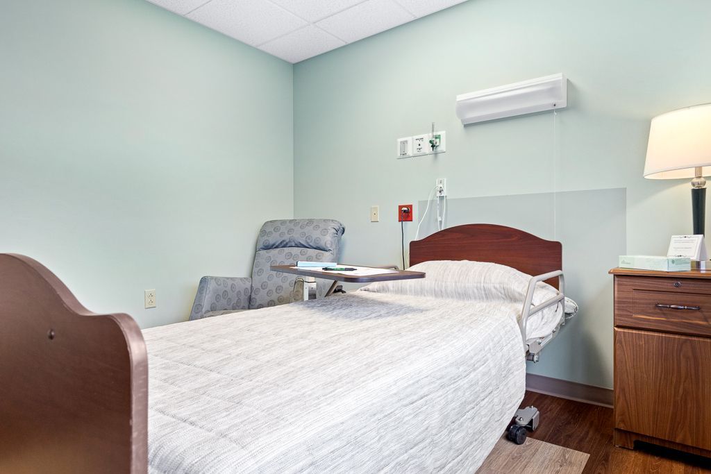 Hospital room with bed, chair, nightstand, and medical equipment. Light green walls, brown bed frame.