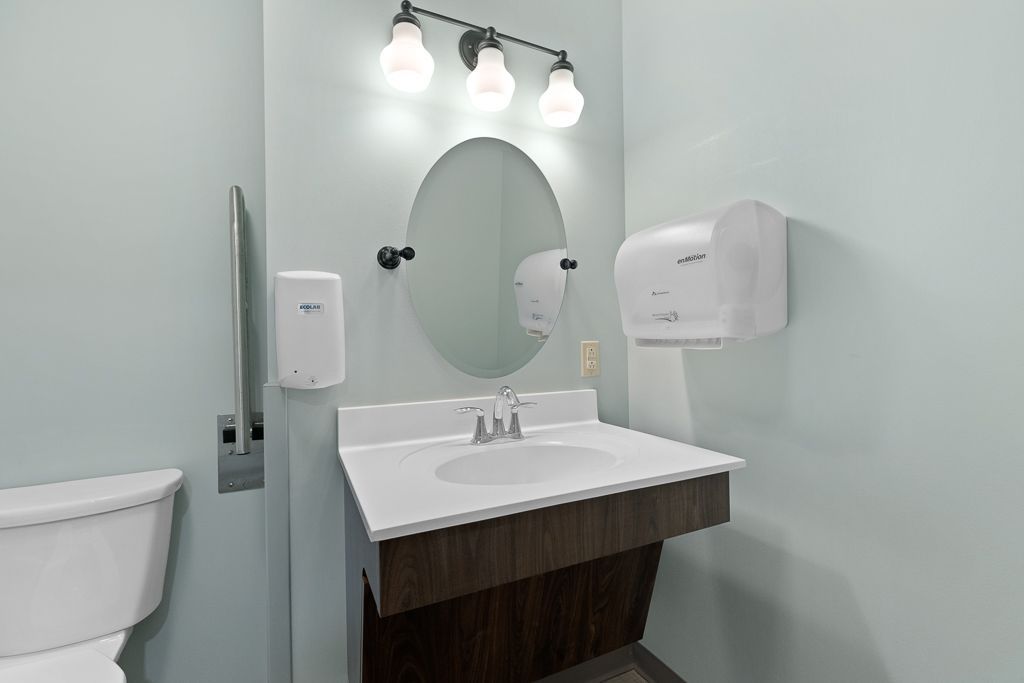 Small bathroom with sink, mirror, and paper towel dispenser. Light blue walls and a white toilet.