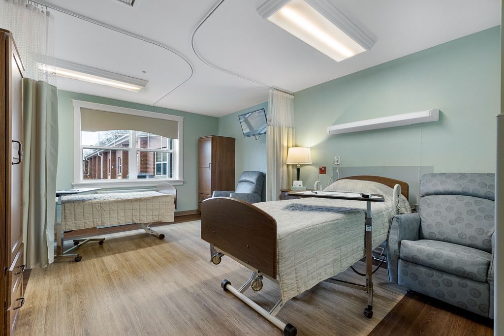 Assisted Living room with adjustable bed, recliner, wardrobe, and window with curtains. Soft blue walls and wood floors.