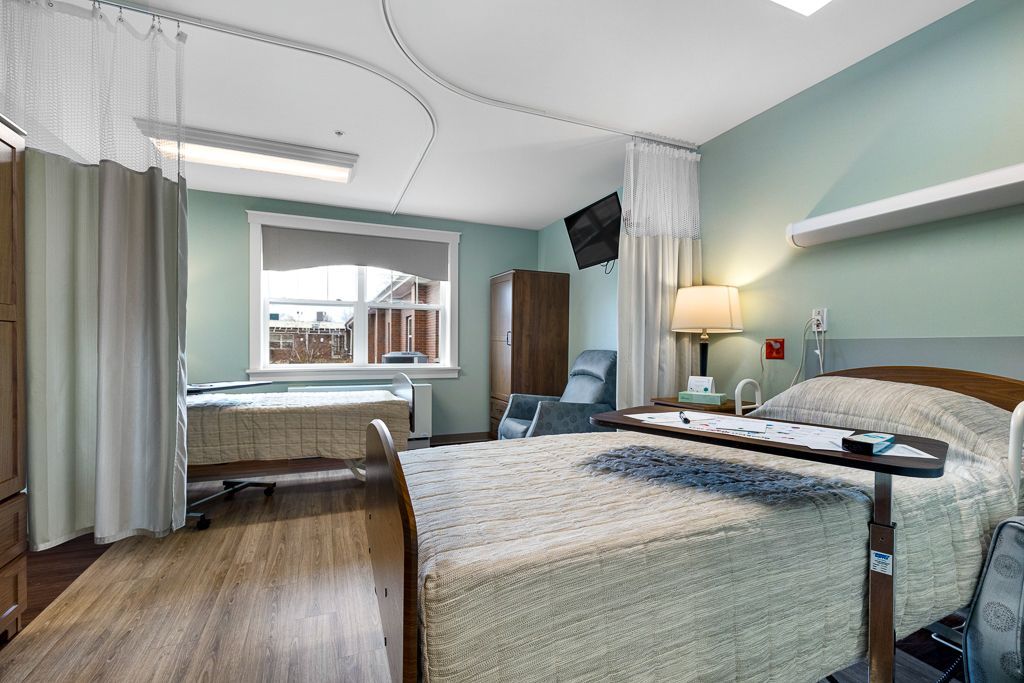 Assisted Living room with two beds, a window, and medical equipment. Pale green walls and wood flooring.