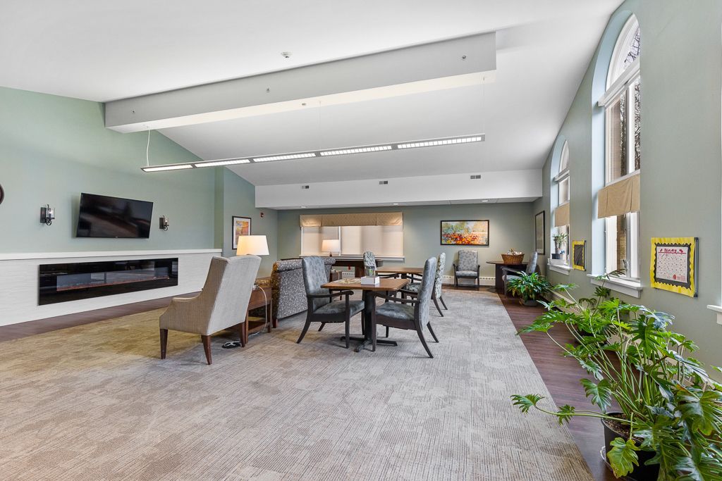 Spacious room with seating areas, TV, and large windows. Light blue walls, light carpet, and plants.