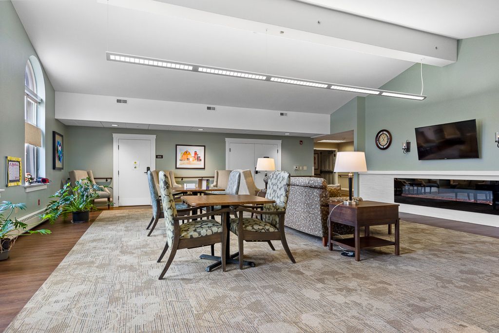 Spacious lounge with tables, chairs, fireplace, and TV in an assisted living facility.