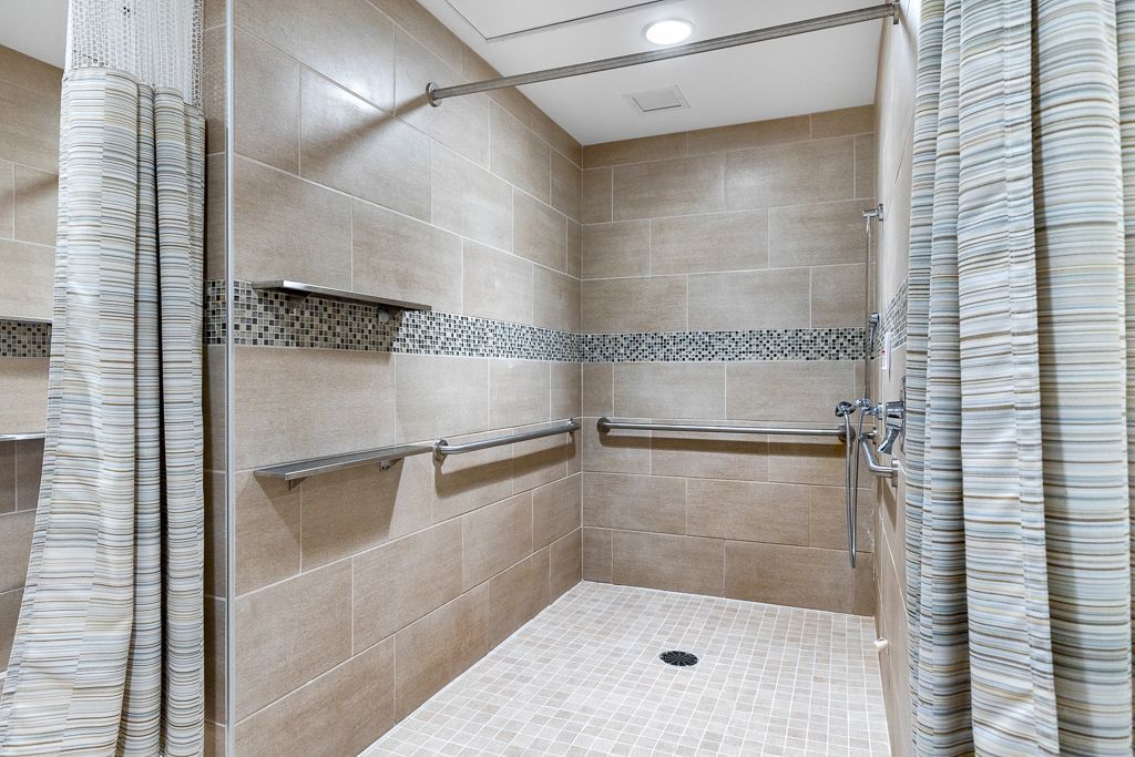 Tile-walled shower with grab bars and a stone-tiled floor.  Beige walls, grey accent strip, shower curtains on sides.