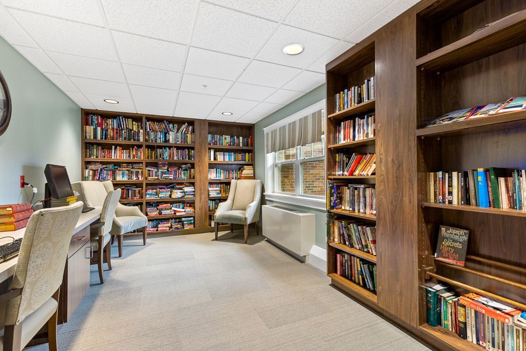 Library with built-in bookshelves filled with books, chairs, desks, and a window. Light-colored carpet and ceiling.