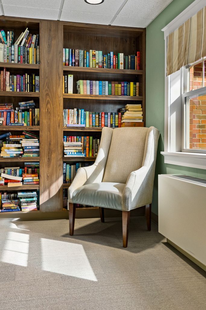 Cozy reading nook: bookshelf, upholstered armchair, window with striped shade, and sunlight.