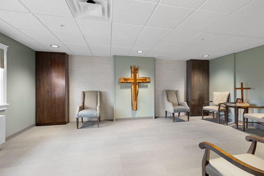 A peaceful room with a large wooden cross on a light green wall, chairs, and two wooden cabinets.