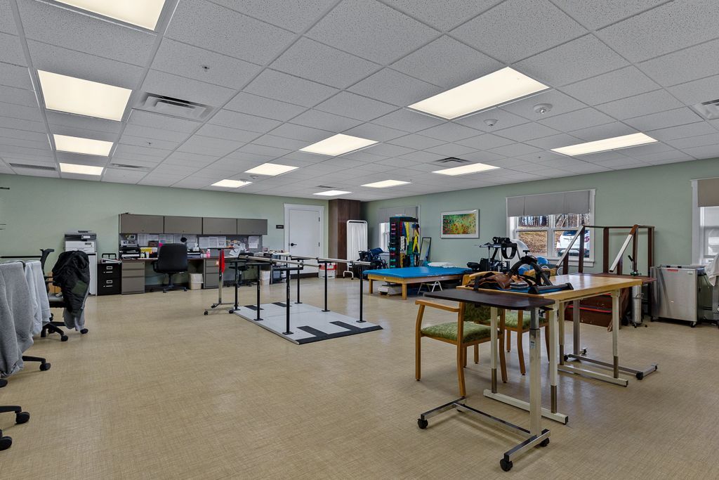Spacious therapy room with tables, equipment, and desks. Bright lighting, neutral walls, and tan flooring.