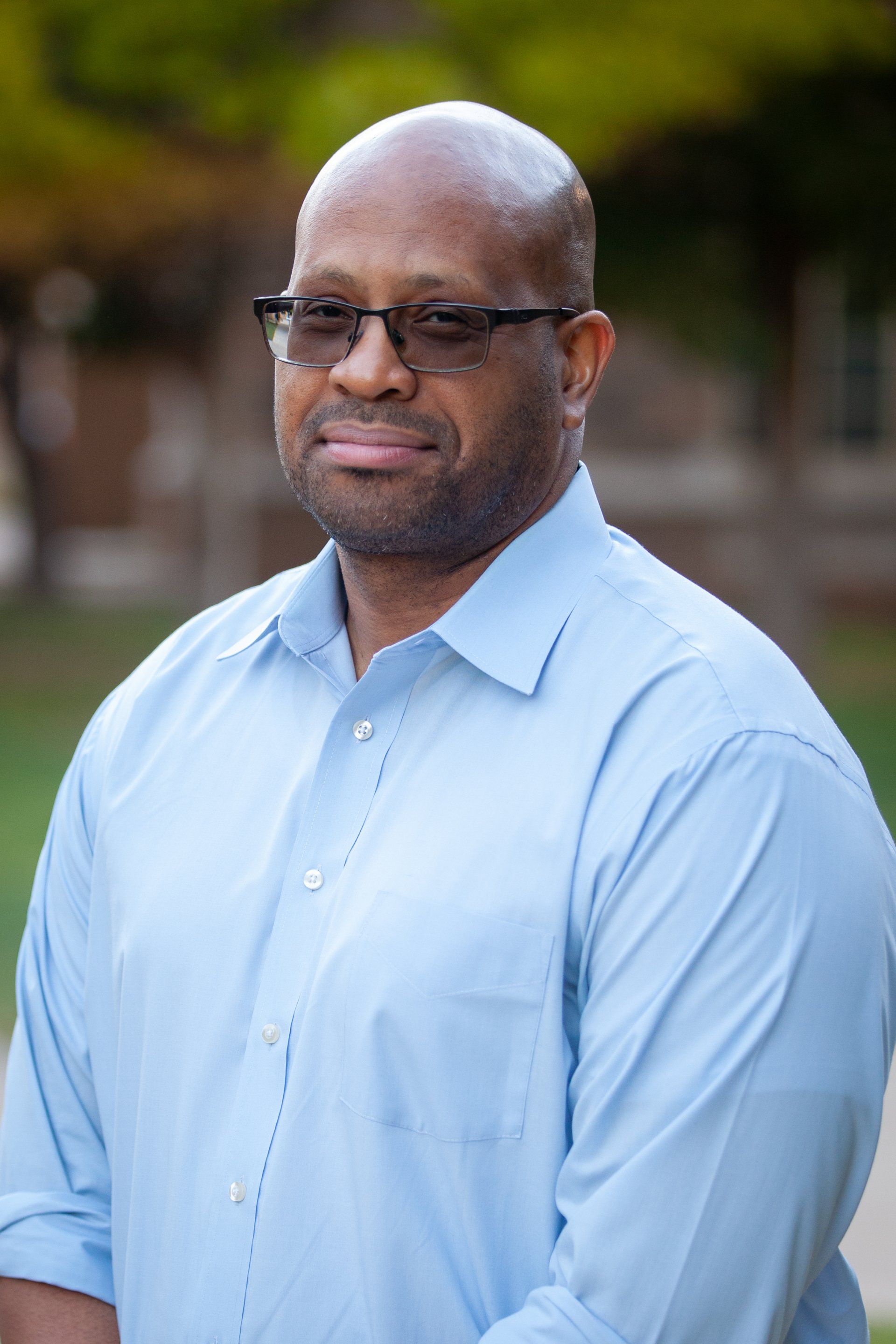 Portrait of a man in a light blue shirt and glasses, standing outdoors with a blurred background