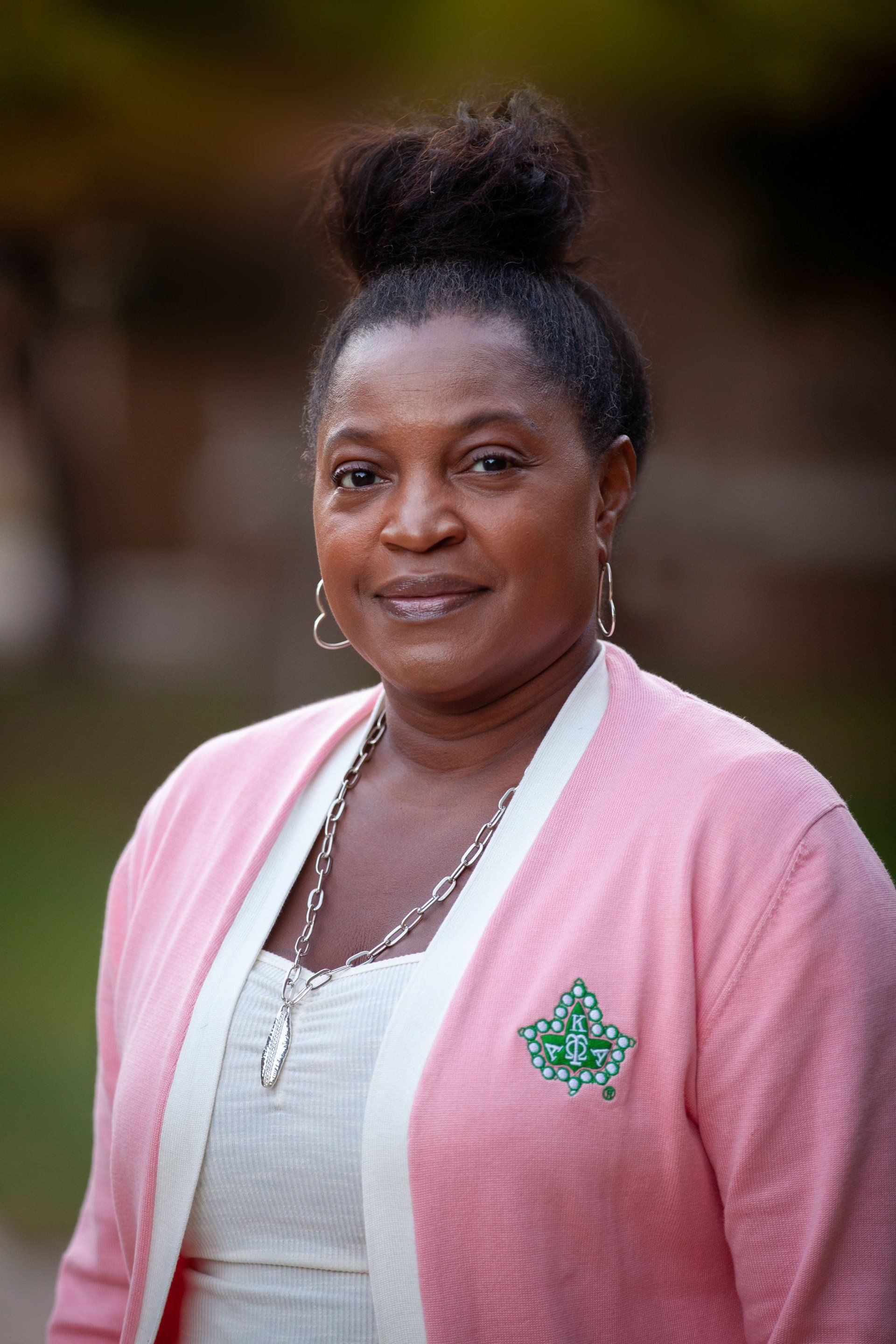 Portrait of a woman in a pink cardigan with a green pin, standing outdoors against a blurred background.