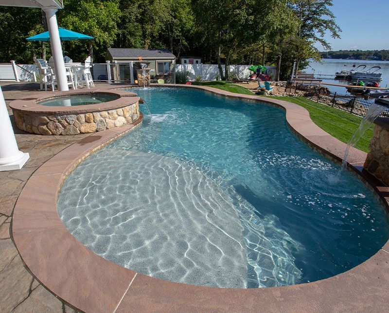 Curved backyard swimming pool with clear blue water, stone edge, and lakeside patio seating