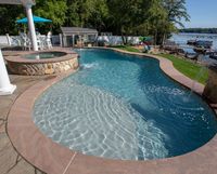 Curved backyard swimming pool with clear blue water, stone edge, and lakeside patio seating