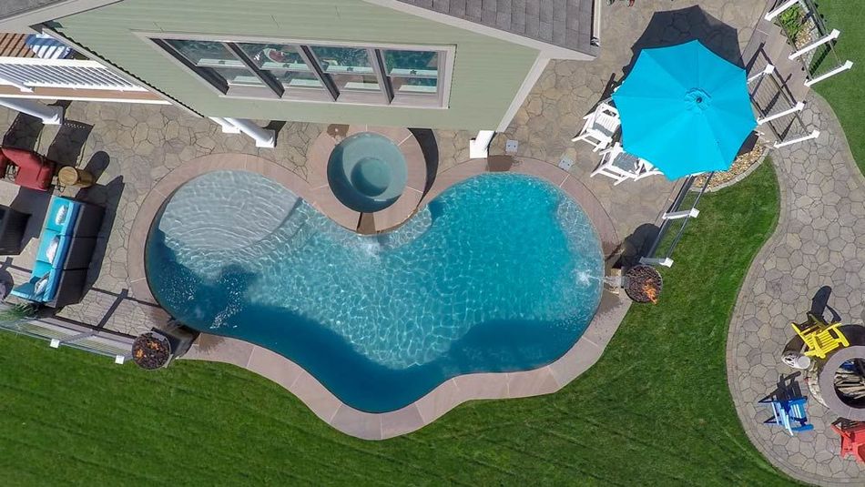 Aerial view of a backyard pool with a hot tub, blue umbrella, lawn, and lounge chairs