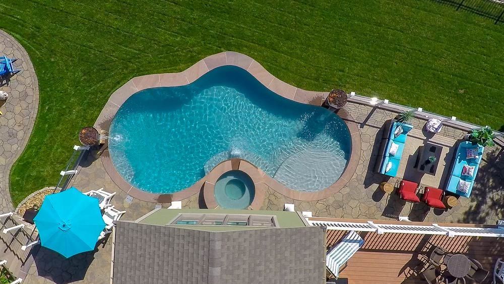 Aerial view of a backyard pool with a hot tub, patio furniture, umbrellas, and green lawn