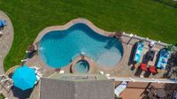 Aerial view of a backyard pool with a hot tub, patio furniture, umbrellas, and green lawn