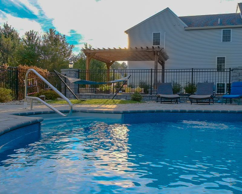 Backyard swimming pool with patio chairs, fence, and a white house at sunset