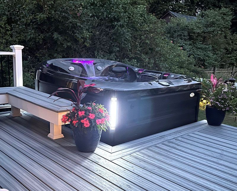 Backyard deck with a black hot tub, lit planters, and a wooden bench at dusk