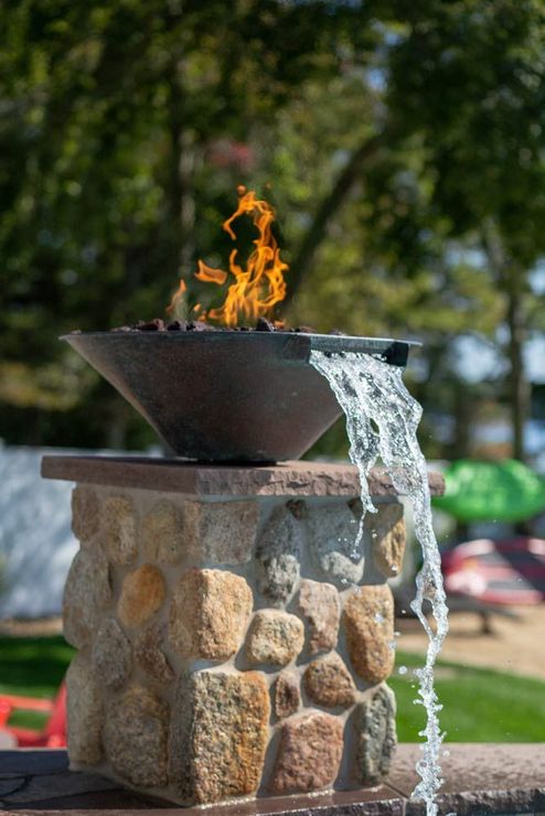 Outdoor stone fountain with a metal basin, flame rising from the top and water spilling over the side.