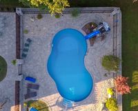Aerial view of a backyard swimming pool with a slide, patio chairs, and surrounding stone paving.