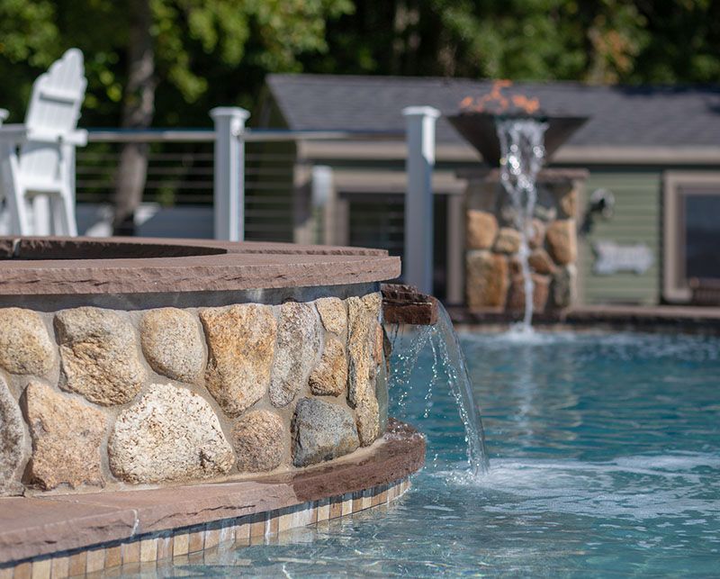 Stone pool edge with water spilling into a blue swimming pool, with a fountain in the background