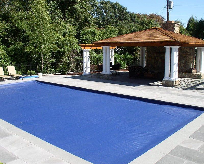 Covered outdoor pool with a bright blue cover beside a stone patio and pavilion