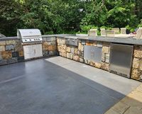 Outdoor stone patio with built-in stainless steel grill and storage cabinets against a low stone wall