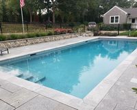 Rectangular outdoor pool with blue water, stone deck, and a fenced backyard with flowers and a small house.