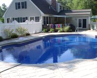 Backyard swimming pool beside a white house with patio chairs and a fence