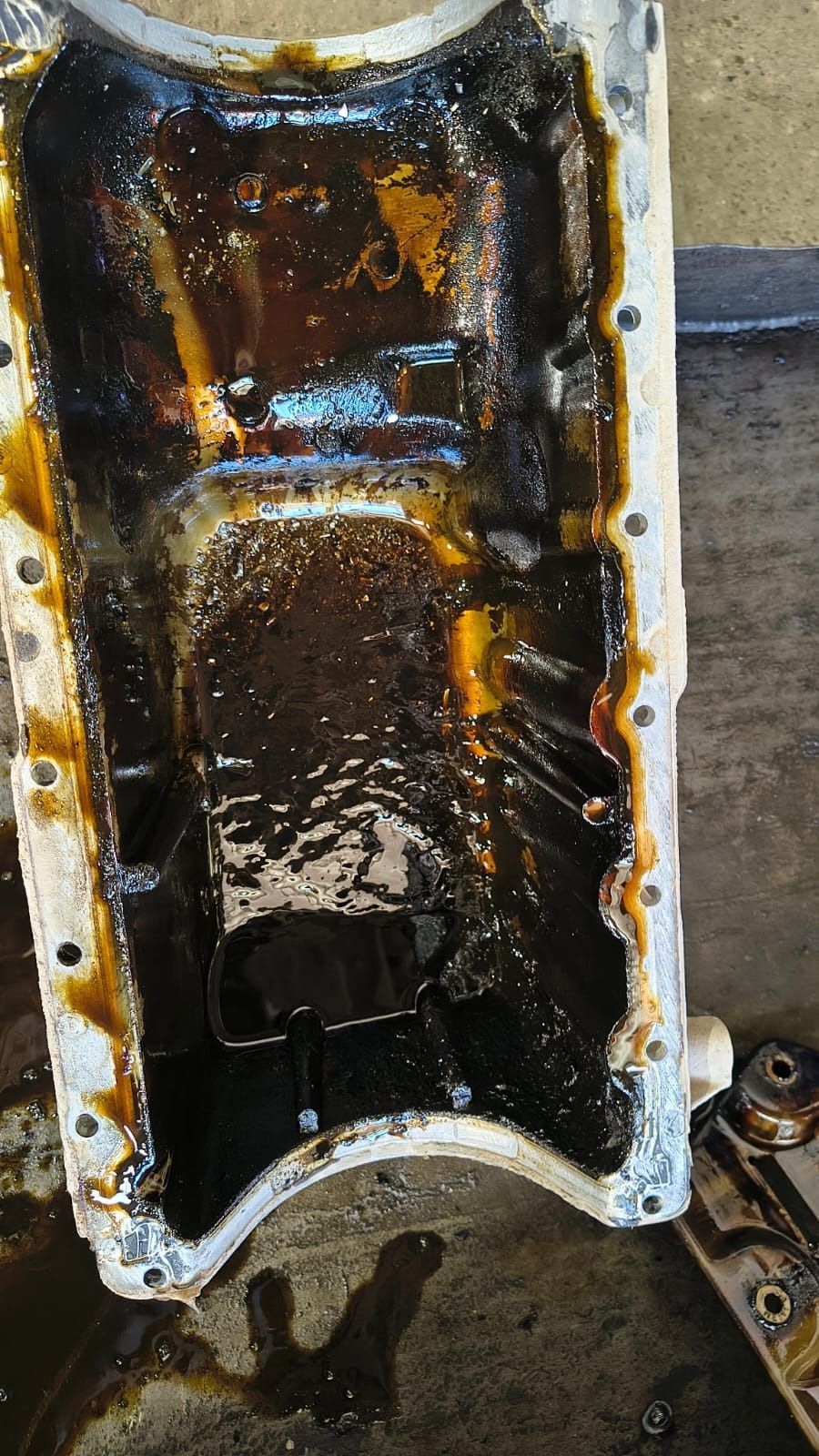 Cárter de motor metálico empapado en aceite con lodo oscuro y residuos en su interior, mostrado sobre una superficie de trabajo.