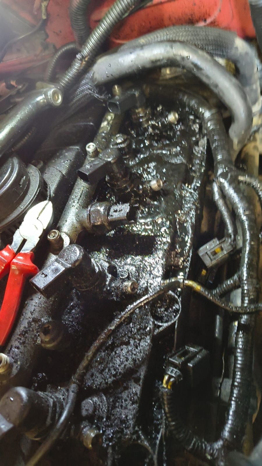 Compartimento del motor desordenado con componentes negros grasientos, mangueras y alicates de mango rojo en primer plano.