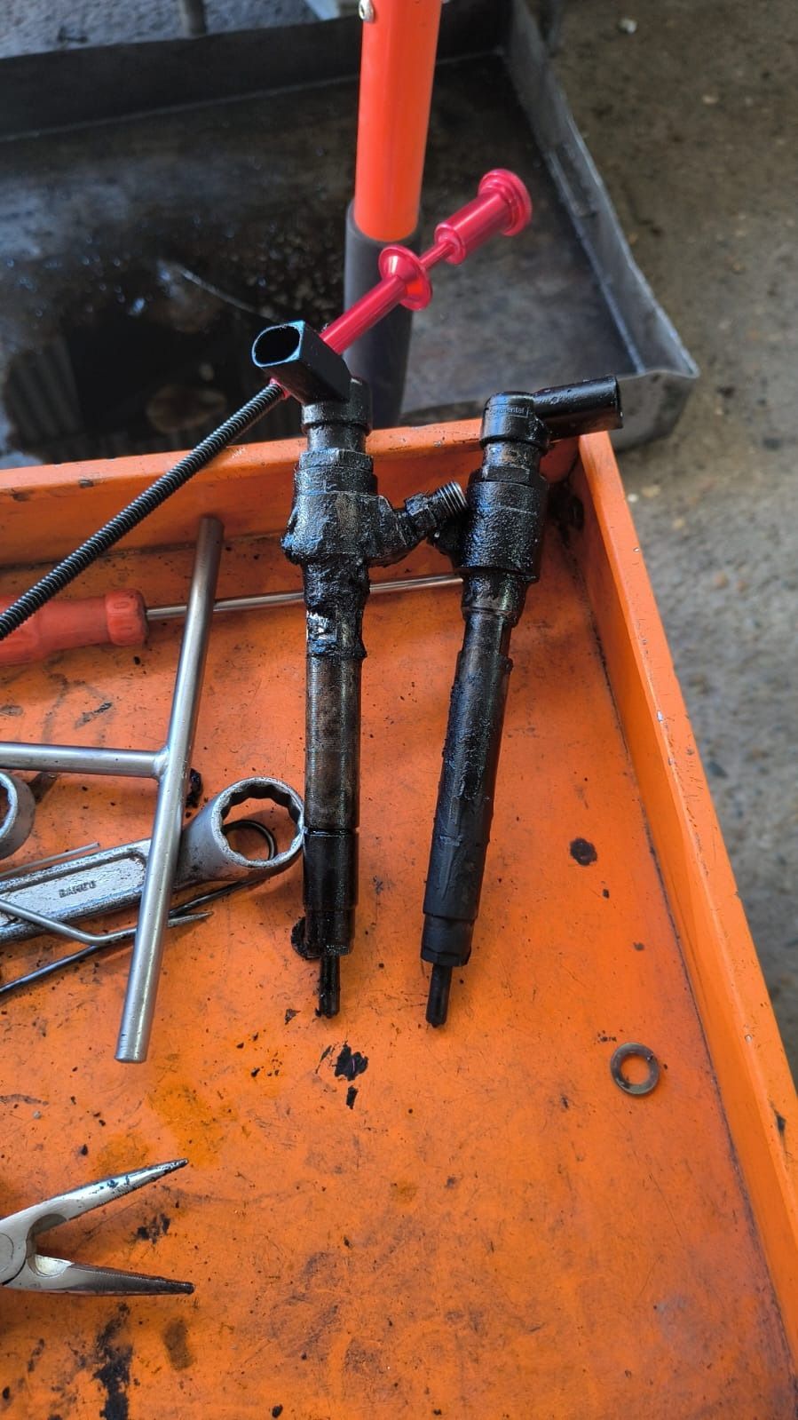 Dos ganchos y herramientas de metal negro y lodoso sobre un banco de trabajo naranja cerca de un mango rojo.