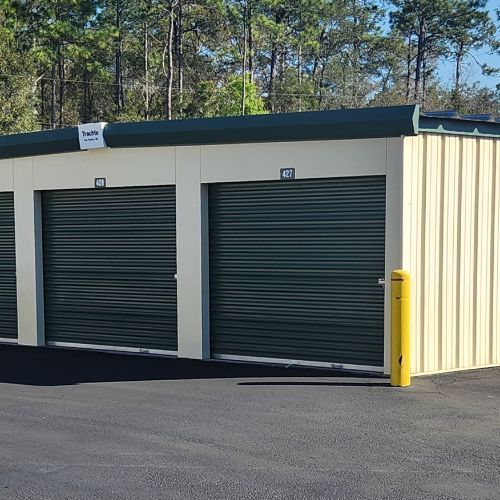 Storage units with green doors and light-yellow walls under a green-roofed structure. Units are in a paved lot with trees in the background.