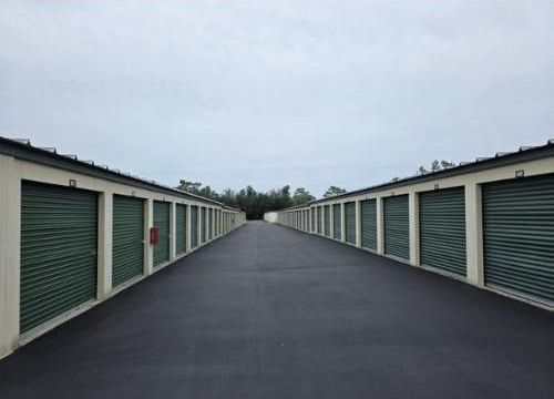 Exterior view of storage units, with dark green doors and light beige walls lining a paved pathway under an overcast sky.