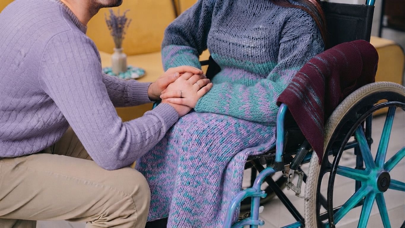 Person in wheelchair being comforted by another person, with a hand on their arm and blanket over their lap.