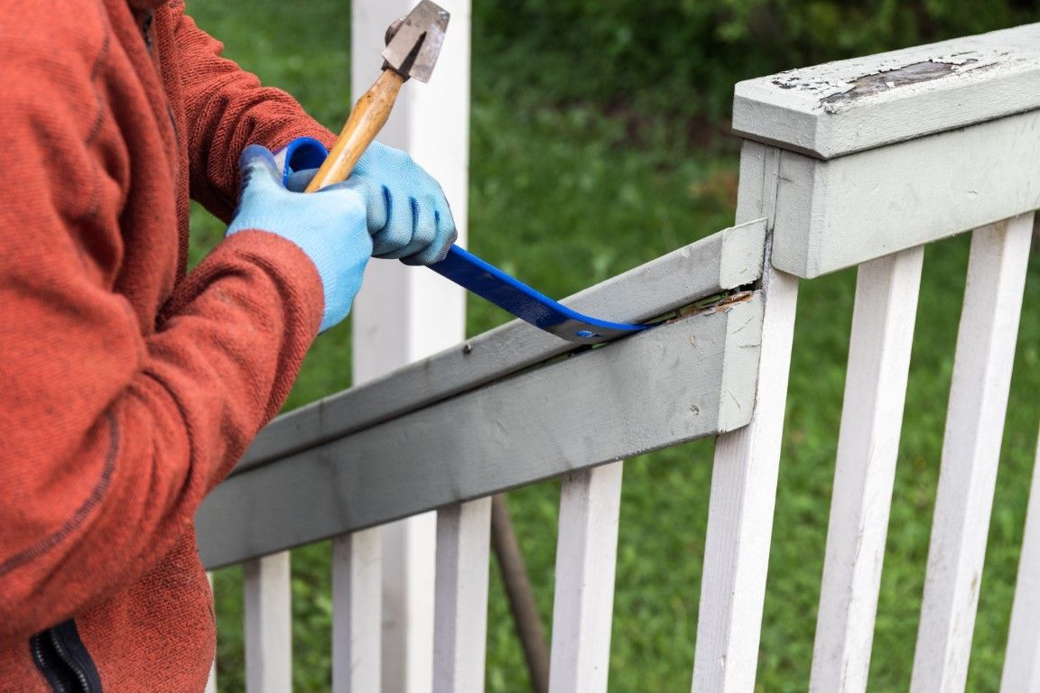 Person painting a white wooden fence with a blue brush and hammer in hand
