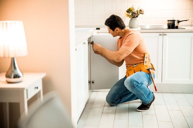 Person kneeling in a kitchen, opening a lower cabinet with a wrench next to a lamp-lit counter.
