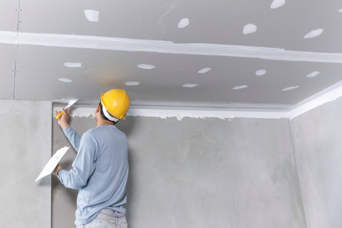 Worker in a yellow hard hat taping drywall seams on a ceiling in a room under construction
