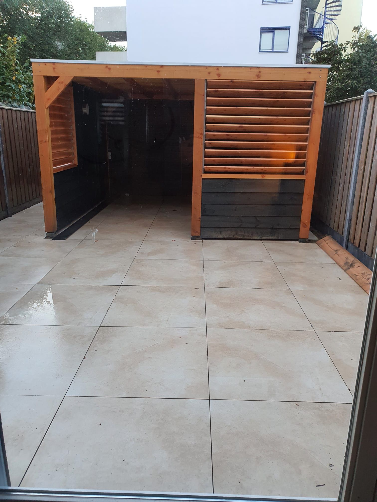 Houten carport of schuur met open voorkant naast een betegeld terras en omheinde tuin.