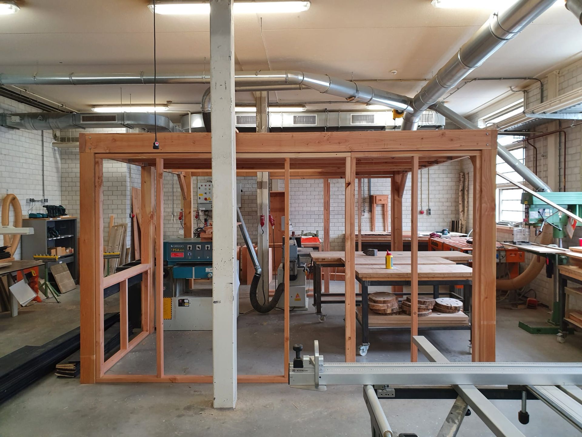 Houten frame in aanbouw in een werkplaats, met gereedschap, werkbanken en een centrale steunpaal.