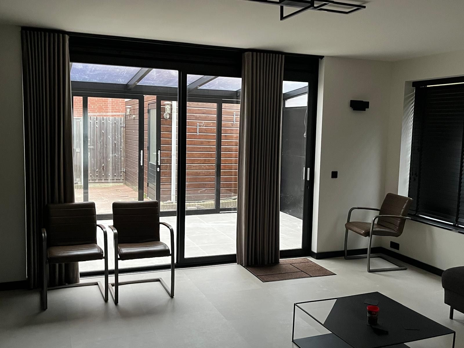 Moderne woonkamer met schuifdeuren van glas, twee stoelen, gordijnen en een kleine salontafel.