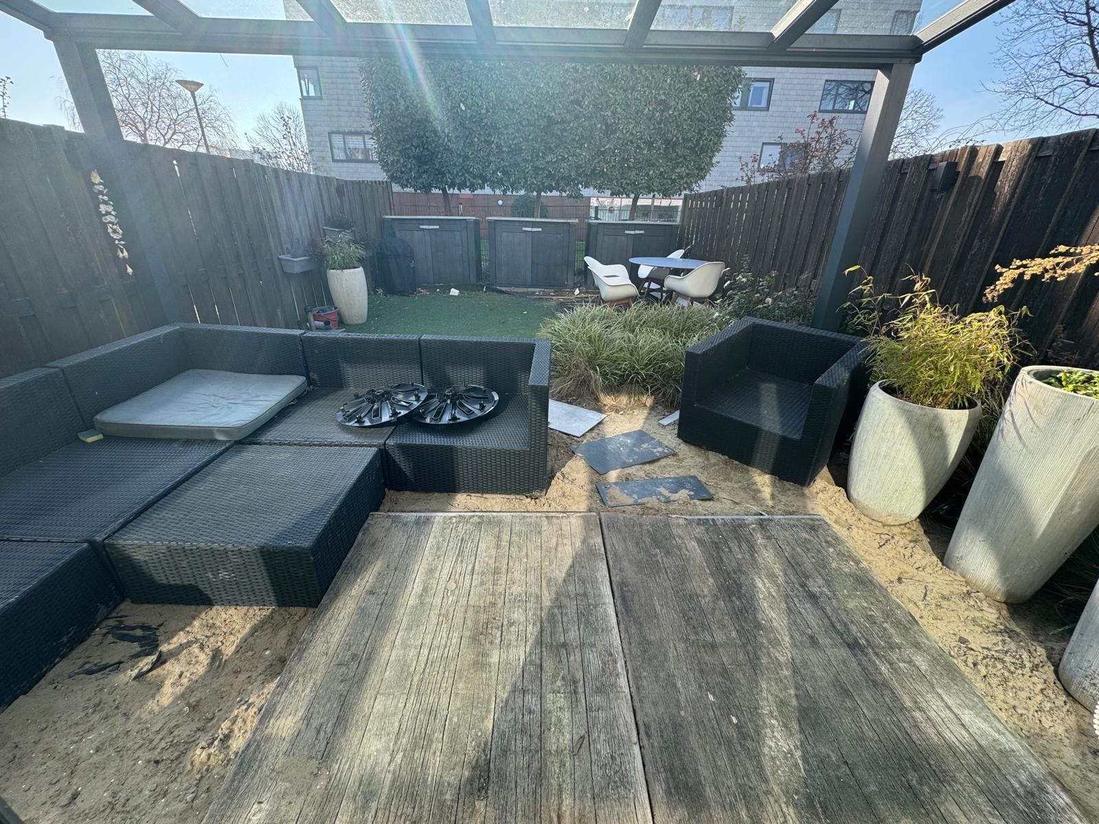 Terras met geweven tuinmeubelen, vuurplaats, plantenbakken en pergola in een zonnige achtertuin.