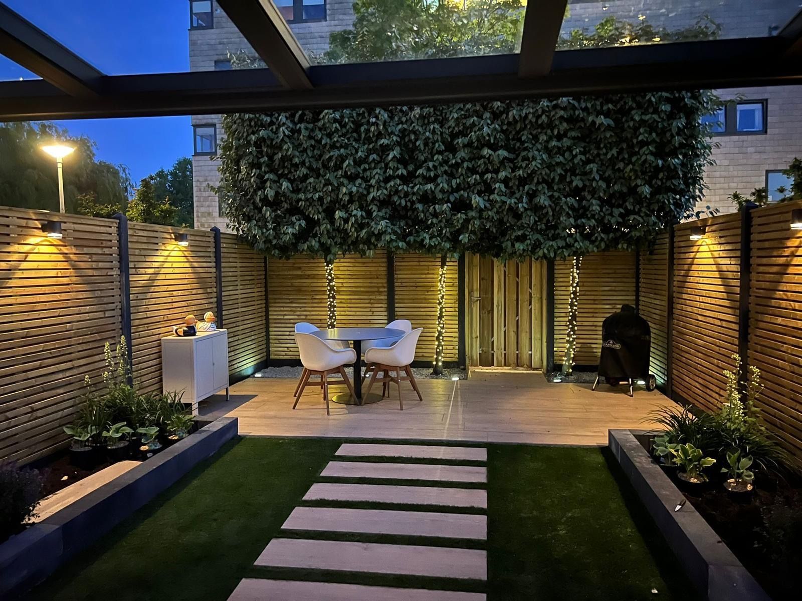 Moderne achtertuin met stenen pad, eettafel, sfeerverlichting en weelderig groen in de schemering.
