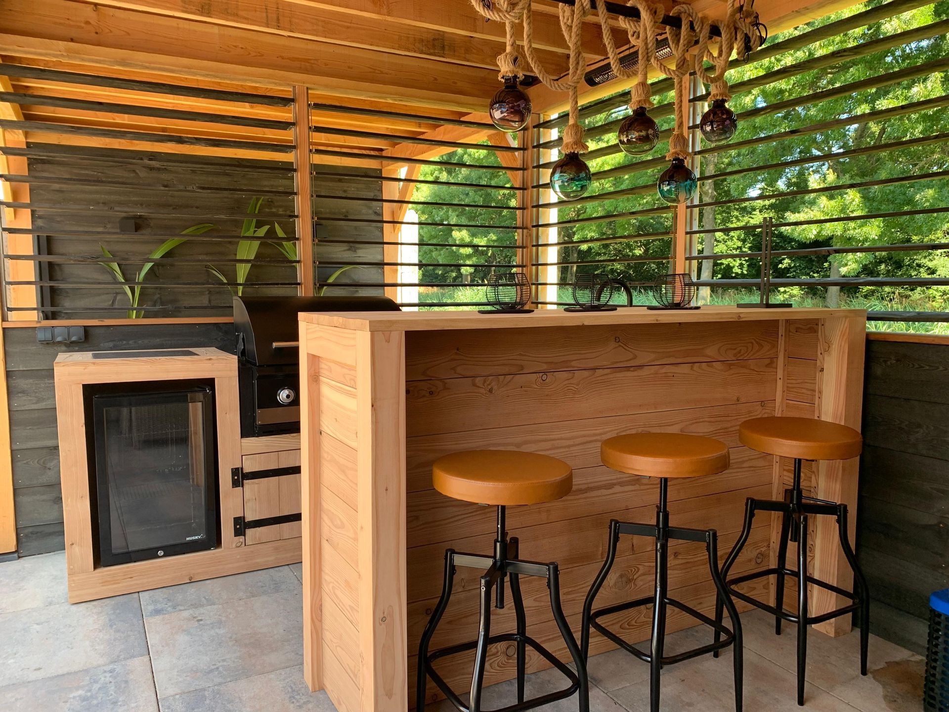 Een houten buitenbar met drie barkrukken op een overdekte veranda, naast een kleine houtkachel en groene begroeiing.