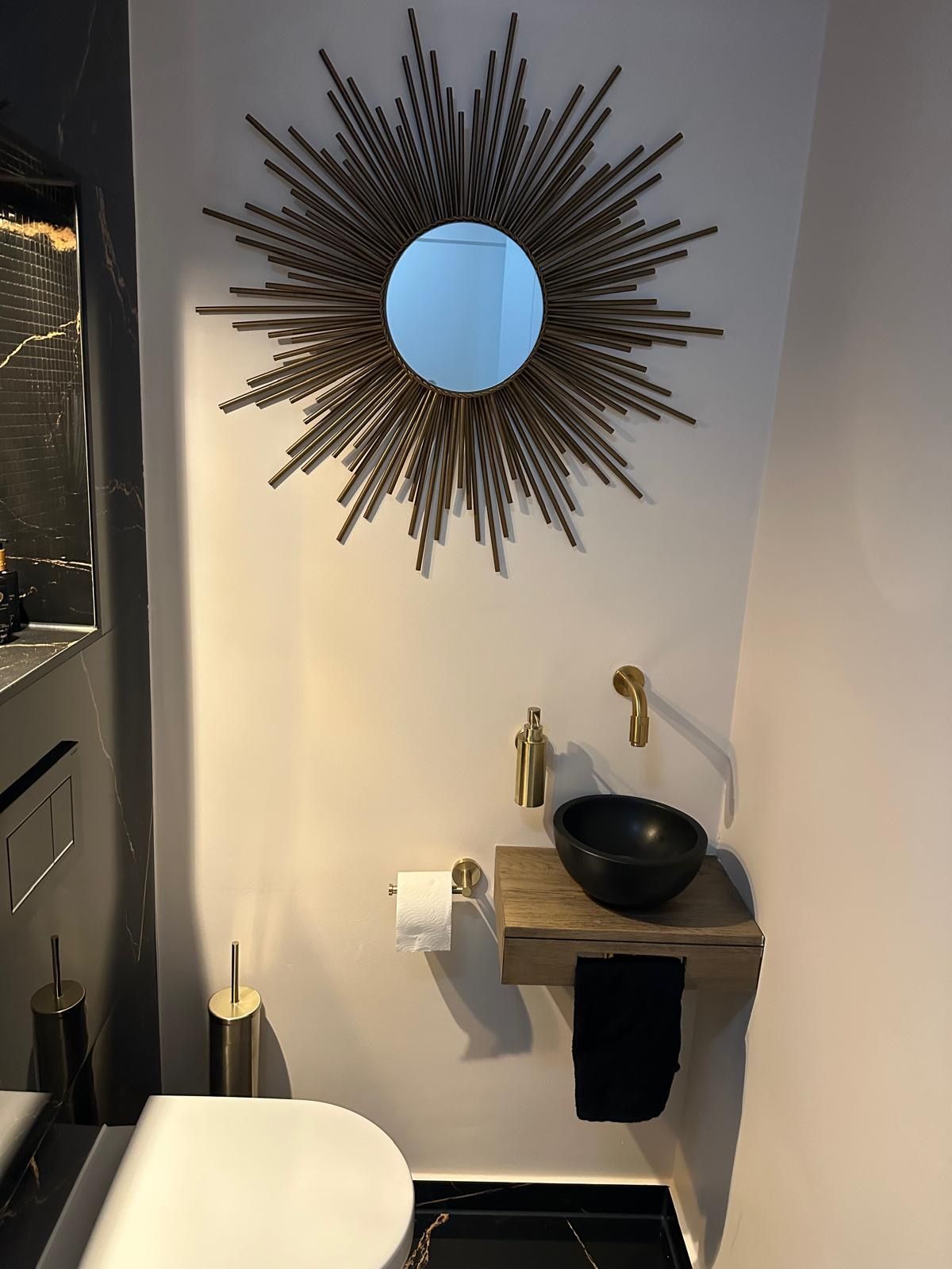 Moderne badkamerkast met ronde spiegel, zwarte wastafel, messing kraan en wanddecoratie met zonnestralenmotief.
