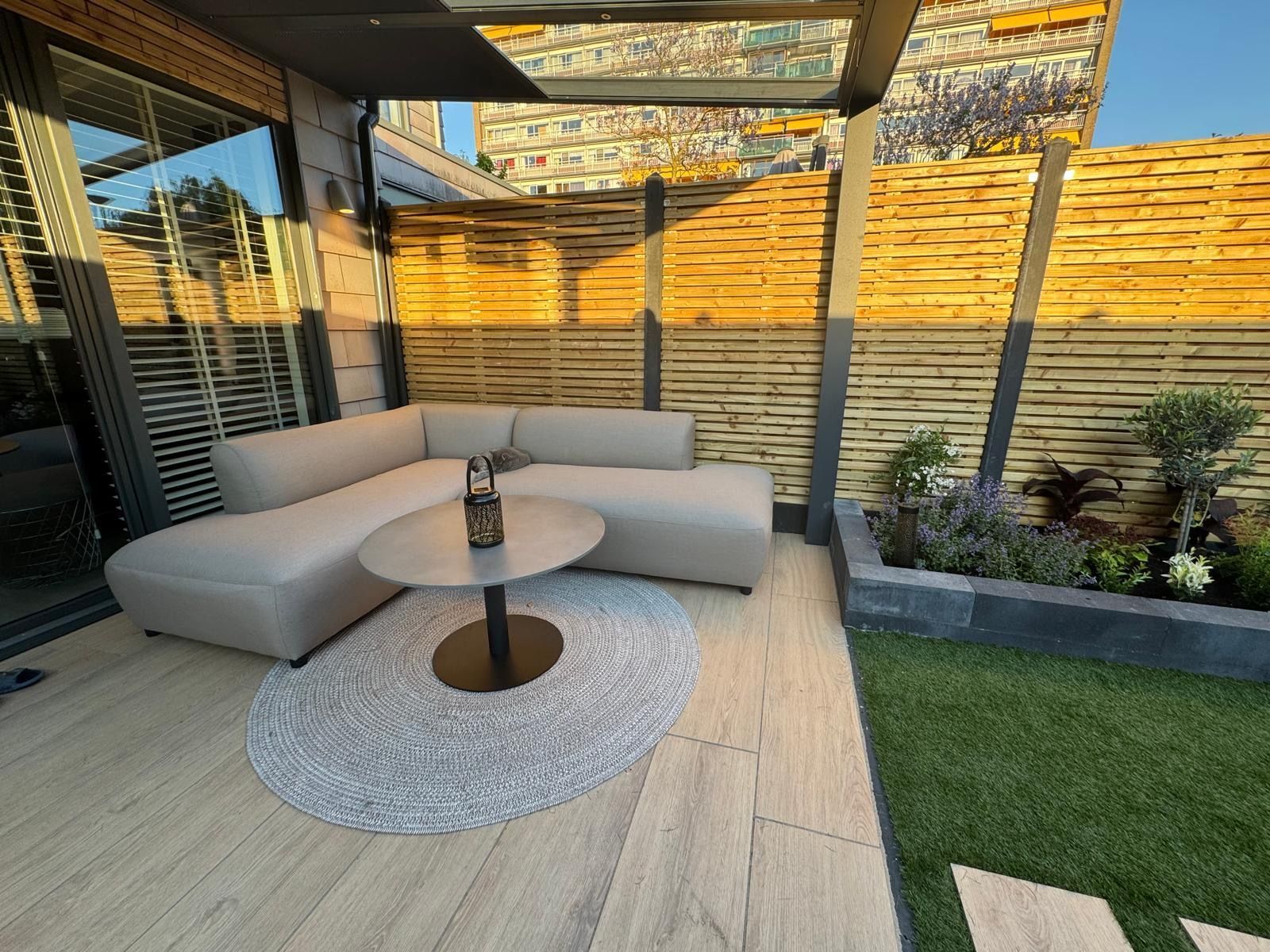 Moderne buitenpatio met beige hoekbank, ronde tafel, geweven vloerkleed en tuinplanten onder een pergola.