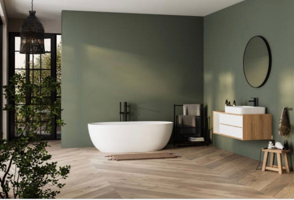 Minimalistische badkamer met een wit bad, groene muren, houten vloer en een ronde spiegel boven een wastafel.