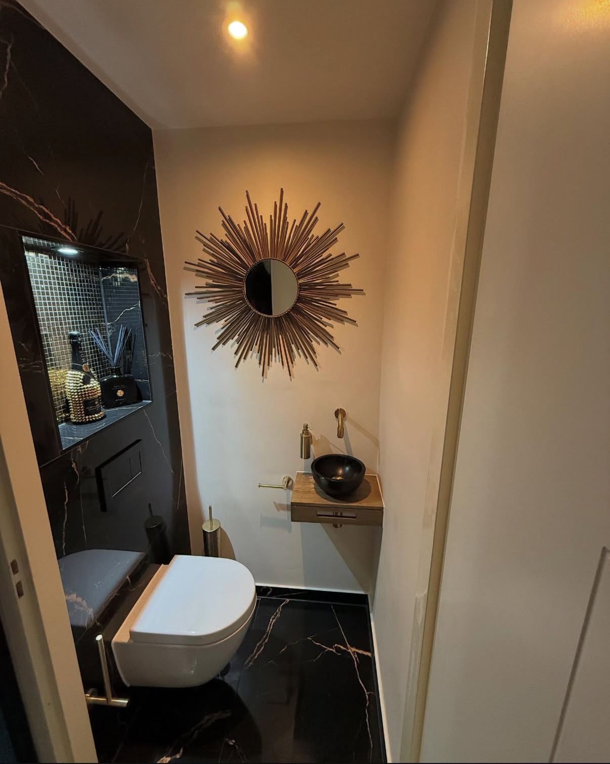 Moderne zwart-witte badkamer met wandgemonteerde wastafel, toilet en gouden spiegel met zonnestralenmotief.