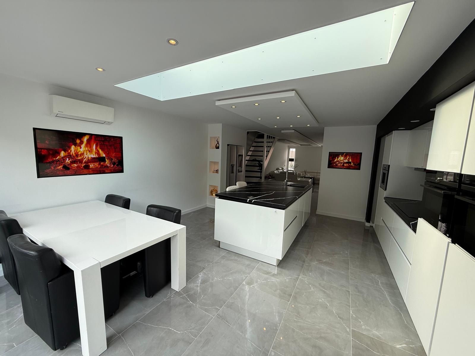 Moderne grijze eetkamer met witte tafel, zwarte stoelen, dakraam en muurdecoratie, die uitkomt op een keuken.