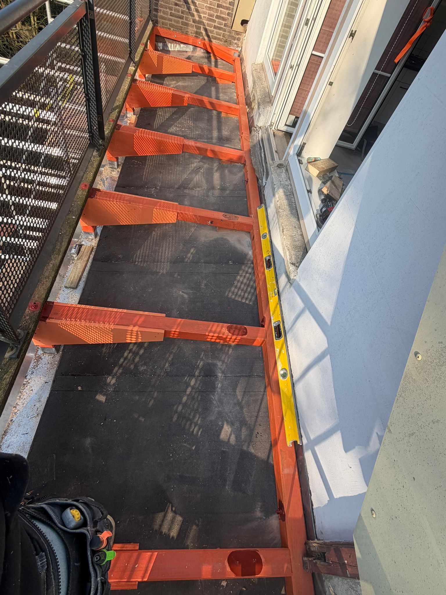 Smalle loopbrug op het dak met oranje veiligheidsnetten en zwarte vloer, langs de muren van het gebouw en een hek.
